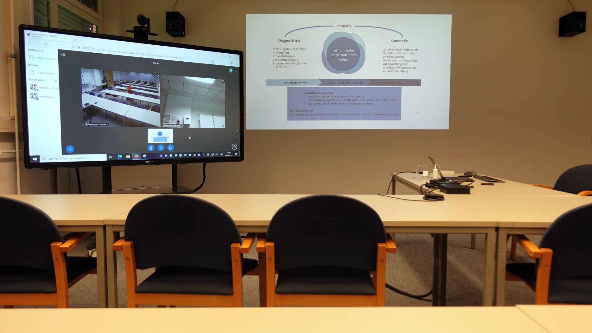 Bild des Seminarraumes mit Sicht aus der zweiten Reihe: Das Display des Konferenzsystems zeigt Webcam-Videos, daneben wird eine Präsentation in Vollformat an die Wand projeziert.